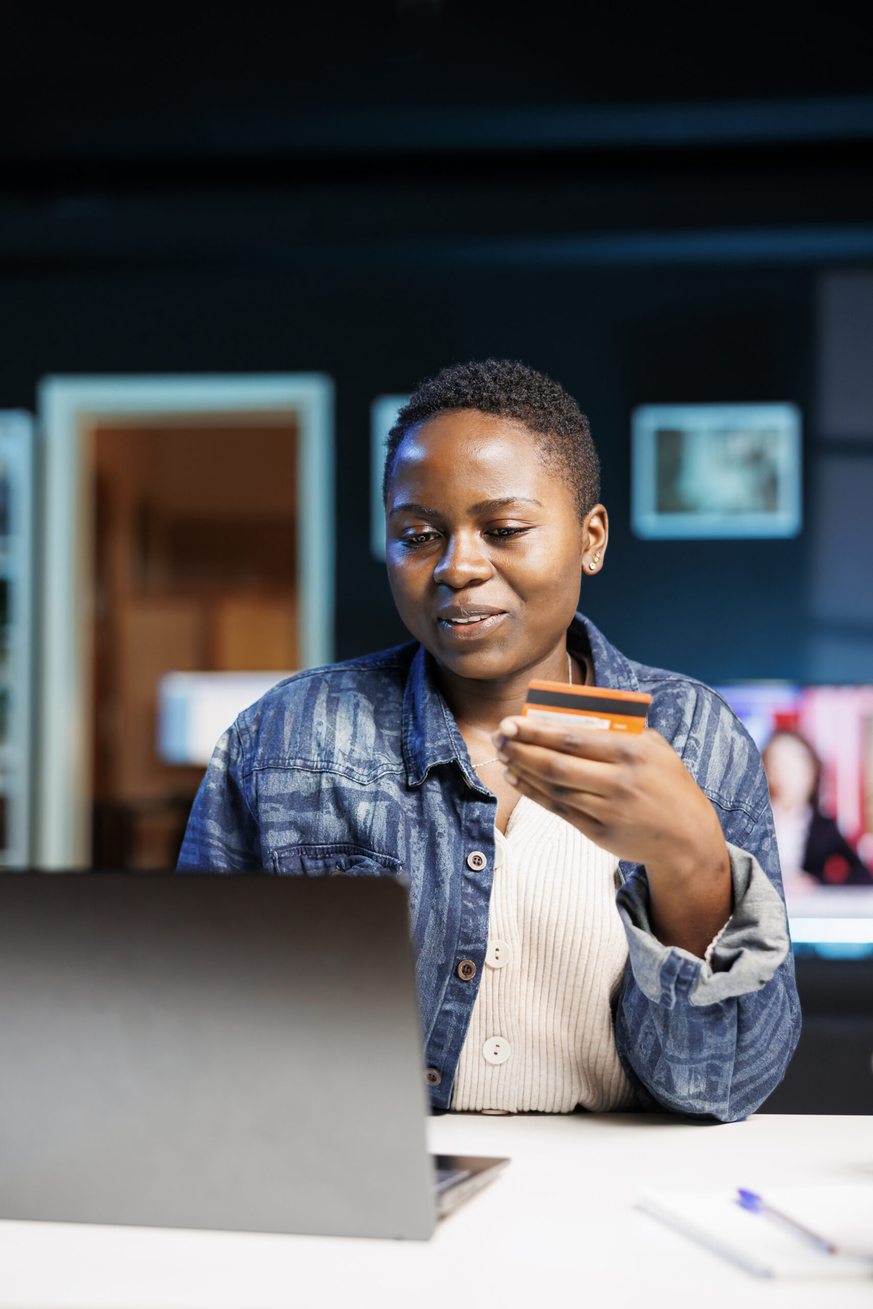 Happy black woman seated at a desk, doing internet shopping on a laptop and paying with a debit card. African American lady making use of credit card, making decisions, and conducting online banking.
